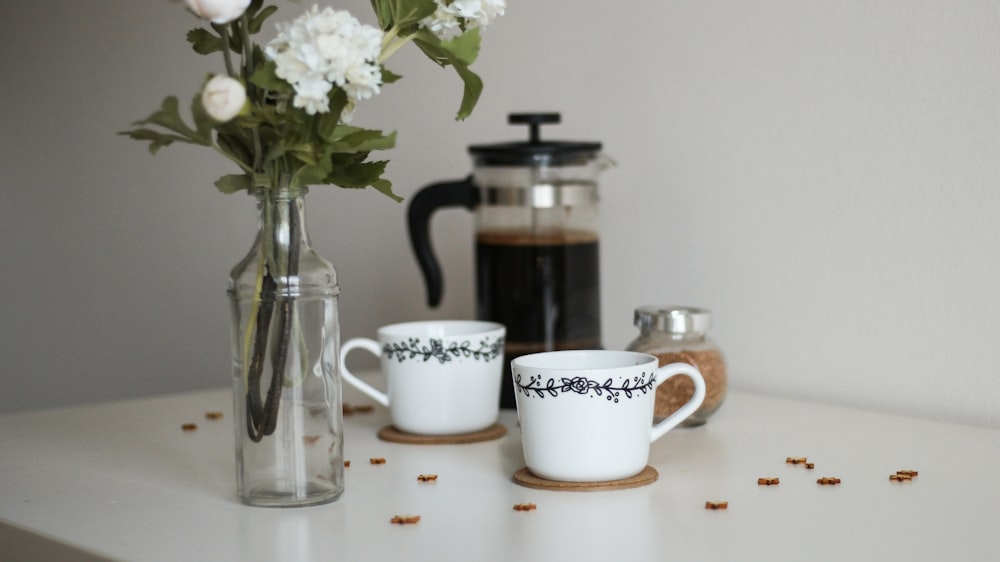 two white ceramic mugs beside clear glass vase and white flowers