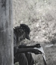 An elderly man with a long beard is sitting outdoors, reading a newspaper. He wears a hat and dark clothing, positioned in a shadowed area with trees or foliage in the blurred background.