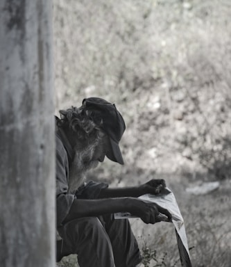 An elderly man with a long beard is sitting outdoors, reading a newspaper. He wears a hat and dark clothing, positioned in a shadowed area with trees or foliage in the blurred background.