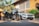 A motorcycle and a luxury car are parked in front of a warm-colored building with architectural features and greenery. The motorcycle is in the foreground, with the white car positioned slightly behind it. Palm trees and plants add a tropical feel to the setting.