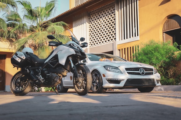 A motorcycle and a luxury car are parked in front of a warm-colored building with architectural features and greenery. The motorcycle is in the foreground, with the white car positioned slightly behind it. Palm trees and plants add a tropical feel to the setting.