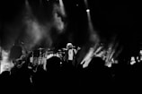 A band performing on stage with dramatic lighting and smoke effects. The lead singer stands in the center, holding a microphone, while other band members play guitars and drums. The audience is partially visible in the foreground, silhouetted against the stage lights.