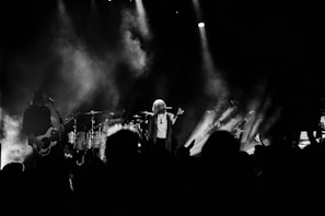 A band performing on stage with dramatic lighting and smoke effects. The lead singer stands in the center, holding a microphone, while other band members play guitars and drums. The audience is partially visible in the foreground, silhouetted against the stage lights.