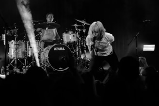 A black and white concert scene with a singer energetically leaning forward with a microphone on stage. A drummer is in the background, playing drums with a focused expression. The audience's heads and hands are visible, indicating an engaged crowd.
