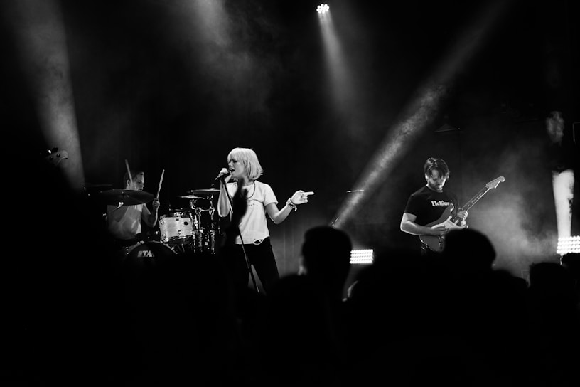 A live band performance featuring a lead singer with short blonde hair holding a microphone. The drummer is energetically playing the drums in the background, while a guitarist strums an electric guitar to the right. The scene is illuminated by stage lights, creating a dynamic and moody atmosphere with smoke effects.