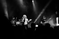 A live band performance featuring a lead singer with short blonde hair holding a microphone. The drummer is energetically playing the drums in the background, while a guitarist strums an electric guitar to the right. The scene is illuminated by stage lights, creating a dynamic and moody atmosphere with smoke effects.