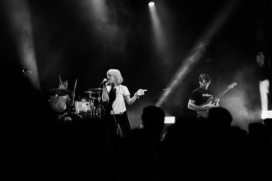 A live band performance featuring a lead singer with short blonde hair holding a microphone. The drummer is energetically playing the drums in the background, while a guitarist strums an electric guitar to the right. The scene is illuminated by stage lights, creating a dynamic and moody atmosphere with smoke effects.