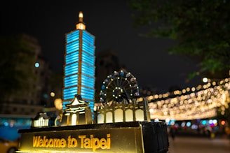 A night scene featuring a miniature model of the Taipei cityscape, including a prominent blue-lit building resembling Taipei 101. The model is adorned with the words 'Welcome to Taipei' in glowing letters. In the background, decorative string lights form a bokeh effect, creating a festive and vibrant atmosphere. A ferris wheel is also part of the miniature display.