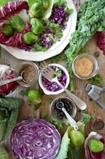 An arrangement of fresh vegetables and whole spices on a rustic wooden surface. There are whole brussels sprouts, chopped purple cabbage, leafy greens, and a halved cabbage head. Small jars hold various seeds and spices, including mustard seeds and black peppercorns, and a metal garlic press is visible.