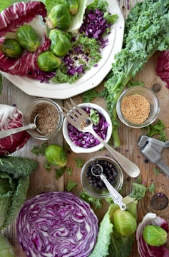 An arrangement of fresh vegetables and whole spices on a rustic wooden surface. There are whole brussels sprouts, chopped purple cabbage, leafy greens, and a halved cabbage head. Small jars hold various seeds and spices, including mustard seeds and black peppercorns, and a metal garlic press is visible.