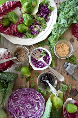 An arrangement of fresh vegetables and whole spices on a rustic wooden surface. There are whole brussels sprouts, chopped purple cabbage, leafy greens, and a halved cabbage head. Small jars hold various seeds and spices, including mustard seeds and black peppercorns, and a metal garlic press is visible.