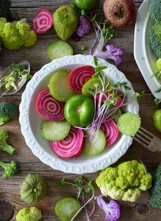 A rustic wooden table features a white plate filled with brightly colored slices of vegetables such as beets and cucumbers, complemented by sprigs of greens. Surrounding the plate are assorted fresh vegetables like broccoli, cauliflower, and tomatillos, creating a vibrant and healthy spread.