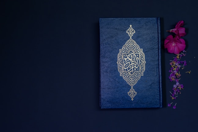 A beautifully adorned book with elaborate gold patterns on a dark blue cover, placed beside vibrant pink and purple flowers against a dark background.