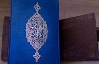 A richly decorated blue book with intricate golden patterns and Arabic script on the cover. The book is resting on a dark brown surface, possibly a wooden table.