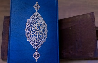 A richly decorated blue book with intricate golden patterns and Arabic script on the cover. The book is resting on a dark brown surface, possibly a wooden table.