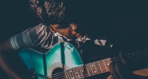 A person is playing a blue acoustic guitar in a dimly lit setting. The light casts dramatic shadows, highlighting the individual's focused expression and the guitar's glossy surface.