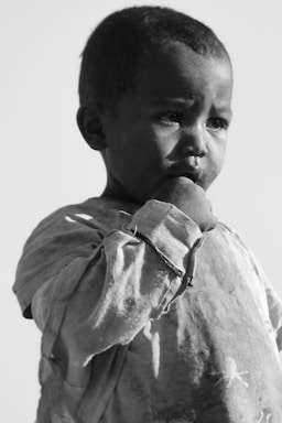 A young child gazes pensively, with a hand near their mouth, wearing a worn, oversized shirt. The monochrome image highlights the contemplative expression and textured fabric.