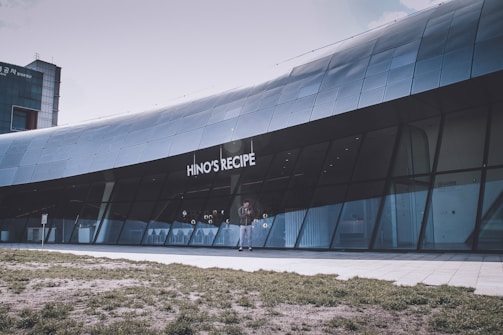 A modern building with a sleek and curved glass facade featuring the name 'HINO'S RECIPE'. The architecture is contemporary, with large windows and metallic panels. In front of the building, a person is walking across a paved and grassy area.