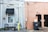 An exterior wall of a building includes a window with a black awning on the left and two black doors on the right. A small seating area with two metal chairs and a table is set up beneath the window, surrounded by gravel. Various utilities, including electrical boxes and cables, are mounted on the wall. A wooden fence section is near the doors, with a garbage bin in front labeled 'Neva.' The building's walls are painted in two different colors: gray and peach.