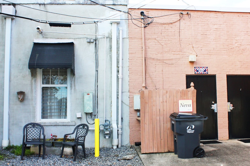 An exterior wall of a building includes a window with a black awning on the left and two black doors on the right. A small seating area with two metal chairs and a table is set up beneath the window, surrounded by gravel. Various utilities, including electrical boxes and cables, are mounted on the wall. A wooden fence section is near the doors, with a garbage bin in front labeled 'Neva.' The building's walls are painted in two different colors: gray and peach.