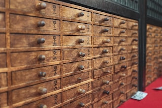 A wooden cabinet with multiple small drawers featuring labels with black text, possibly in a traditional Asian script. Each drawer has a small round handle. The wood appears aged, contributing to a rustic and antique aesthetic.