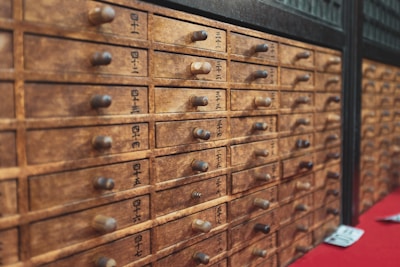 A wooden cabinet with multiple small drawers featuring labels with black text, possibly in a traditional Asian script. Each drawer has a small round handle. The wood appears aged, contributing to a rustic and antique aesthetic.