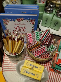 A selection of decorative items including Christmas-themed ornaments resembling butter sticks, cookies, and crackers are displayed. A series of cookbooks titled 'Betty Crocker Lost Recipes' are stacked in the background. An assortment of kitchenware, including salt and pepper shakers, is also visible.