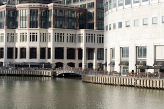 Several modern buildings with large windows are situated along a waterfront. The structures have multiple stories and feature a combination of white and brown exteriors. A promenade runs along the edge of the water, with outdoor seating areas and a pedestrian bridge crossing over. The scene is calm with clear reflections on the water.