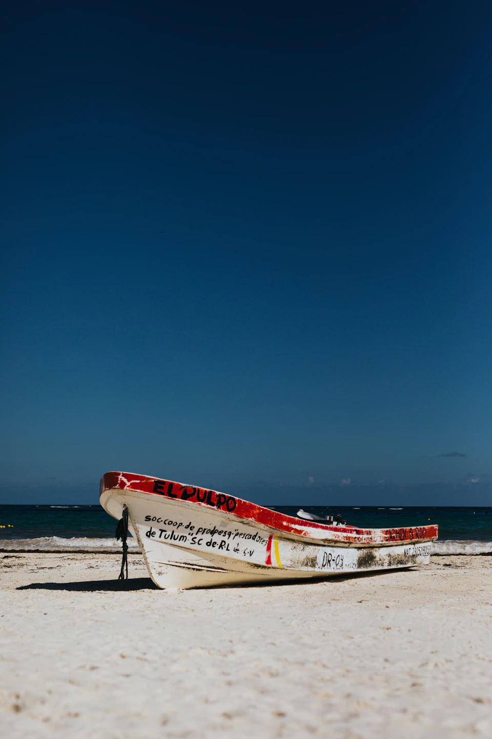 The image depicts a white boat with red accents, docked on a sandy beach under a clear blue sky. The boat is positioned towards the left side of the frame and features a sign that reads "ED'S PORTO". In the background, there are other boats visible in the distance, suggesting this could be a popular spot for boating activities or a harbor area.

The perspective of the image is from ground level, looking up at the boat, which gives it an imposing and majestic appearance. The contrast between the white boat and the blue sky adds to its visual appeal.