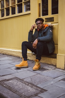 A person sits on a ledge outside a building with yellow walls and windows. They are looking at the camera, holding a cellphone to their ear with one hand and resting the other hand on their knee. The person is wearing a dark green jacket with a fur-lined hood, black jeans, and tan work boots.