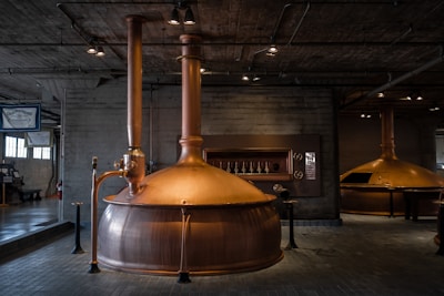 A rustic brewing facility with large copper brewing vats is situated in an industrial-style room. The ceiling features exposed piping and lighting. The walls and floor are made of gray concrete, enhancing the industrial ambiance. Signage is visible in the background, suggesting the facility's identity.