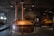 A rustic brewing facility with large copper brewing vats is situated in an industrial-style room. The ceiling features exposed piping and lighting. The walls and floor are made of gray concrete, enhancing the industrial ambiance. Signage is visible in the background, suggesting the facility's identity.