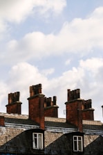 Several brick chimneys rise above a row of traditional slate-roofed buildings. The chimneys vary in size and are topped with multiple clay chimney pots. The sky is filled with fluffy, white clouds against a pale blue backdrop.