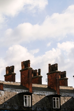 Several brick chimneys rise above a row of traditional slate-roofed buildings. The chimneys vary in size and are topped with multiple clay chimney pots. The sky is filled with fluffy, white clouds against a pale blue backdrop.