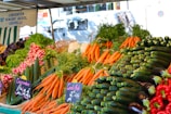 A vibrant market stall displays a colorful array of fresh vegetables including carrots, cucumbers, radishes, red peppers, and eggplants. Lush bunches of parsley and lettuce add greenery. Two chalkboard signs denote prices in French. The background features a striped awning and a blurry street scene.