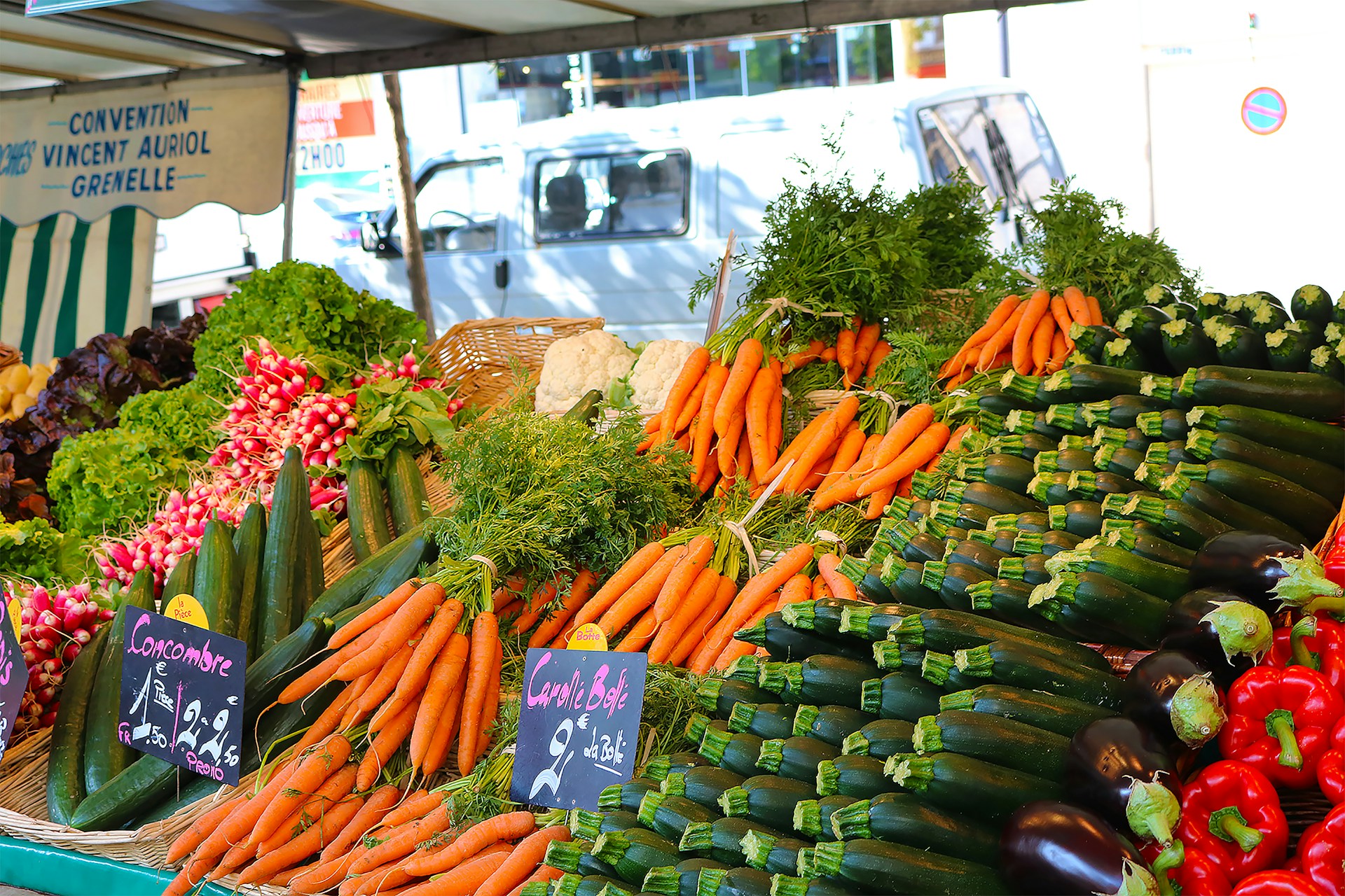 A vibrant market stall displays a colorful array of fresh vegetables including carrots, cucumbers, radishes, red peppers, and eggplants. Lush bunches of parsley and lettuce add greenery. Two chalkboard signs denote prices in French. The background features a striped awning and a blurry street scene.