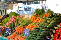 A vibrant market stall displays a colorful array of fresh vegetables including carrots, cucumbers, radishes, red peppers, and eggplants. Lush bunches of parsley and lettuce add greenery. Two chalkboard signs denote prices in French. The background features a striped awning and a blurry street scene.