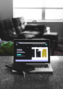 A laptop displaying a software development interface is placed on a granite countertop. Next to the laptop is a closed notebook and a pen. In the background, a sofa and a large window can be seen, providing a contrast between the focused workstation and the casual home setting.