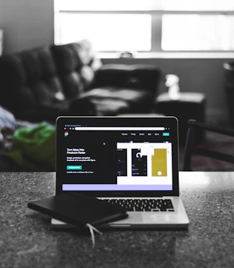 A laptop displaying a software development interface is placed on a granite countertop. Next to the laptop is a closed notebook and a pen. In the background, a sofa and a large window can be seen, providing a contrast between the focused workstation and the casual home setting.