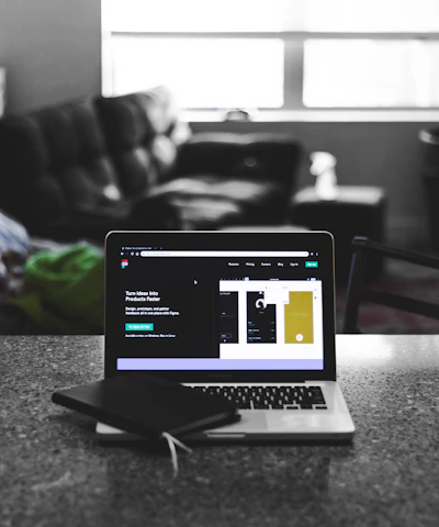 A laptop displaying a software development interface is placed on a granite countertop. Next to the laptop is a closed notebook and a pen. In the background, a sofa and a large window can be seen, providing a contrast between the focused workstation and the casual home setting.