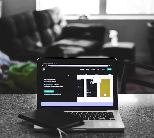A laptop displaying a software development interface is placed on a granite countertop. Next to the laptop is a closed notebook and a pen. In the background, a sofa and a large window can be seen, providing a contrast between the focused workstation and the casual home setting.
