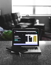 A laptop displaying a software development interface is placed on a granite countertop. Next to the laptop is a closed notebook and a pen. In the background, a sofa and a large window can be seen, providing a contrast between the focused workstation and the casual home setting.