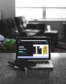 A laptop displaying a software development interface is placed on a granite countertop. Next to the laptop is a closed notebook and a pen. In the background, a sofa and a large window can be seen, providing a contrast between the focused workstation and the casual home setting.