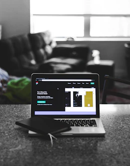 A laptop displaying a software development interface is placed on a granite countertop. Next to the laptop is a closed notebook and a pen. In the background, a sofa and a large window can be seen, providing a contrast between the focused workstation and the casual home setting.