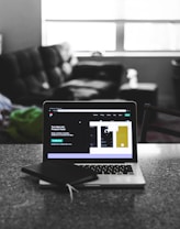 A laptop displaying a software development interface is placed on a granite countertop. Next to the laptop is a closed notebook and a pen. In the background, a sofa and a large window can be seen, providing a contrast between the focused workstation and the casual home setting.