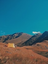 A peaceful mountainous landscape with rolling hills and a vibrant blue sky. A single yellow tent is pitched on the brown, grassy terrain, suggesting an outdoor camping experience. The mountains are rugged and create a stunning backdrop.