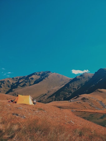 A peaceful mountainous landscape with rolling hills and a vibrant blue sky. A single yellow tent is pitched on the brown, grassy terrain, suggesting an outdoor camping experience. The mountains are rugged and create a stunning backdrop.