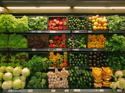 A variety of fresh vegetables are neatly arranged on multiple shelves in a grocery store. The assortment includes leafy greens, red and yellow bell peppers, zucchinis, yellow squash, eggplants, and cauliflower. The produce is organized in rows with similar items grouped together, showcasing vibrant colors and freshness under the store's lighting.
