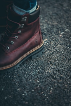 A close-up of a brown leather boot with metal eyelets and sturdy laces on a rough asphalt surface. The boot has thick, rugged soles, and a small brand logo is embossed on the side.