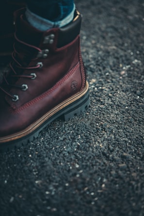 A close-up of a brown leather boot with metal eyelets and sturdy laces on a rough asphalt surface. The boot has thick, rugged soles, and a small brand logo is embossed on the side.